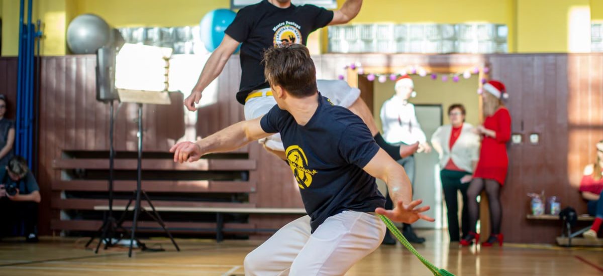 Energetic capoeira performance indoors with audience during daytime.